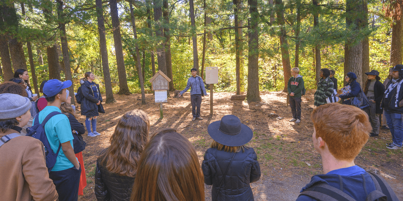 A photograph of a group of people standing in a half-circle in a wooded area. One person stands beside a wooden placard. 