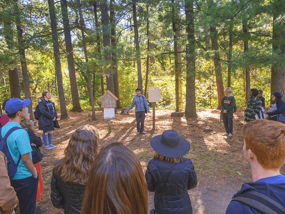 A photograph of a group of people standing in a half-circle in a wooded area. One person stands beside a wooden placard. 