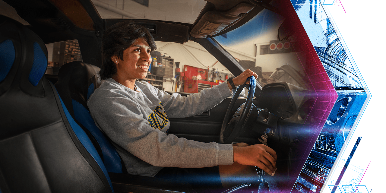 In a photograph, a student wearing a gray University of Michigan crewneck sits in the driver’s seat of a car. 