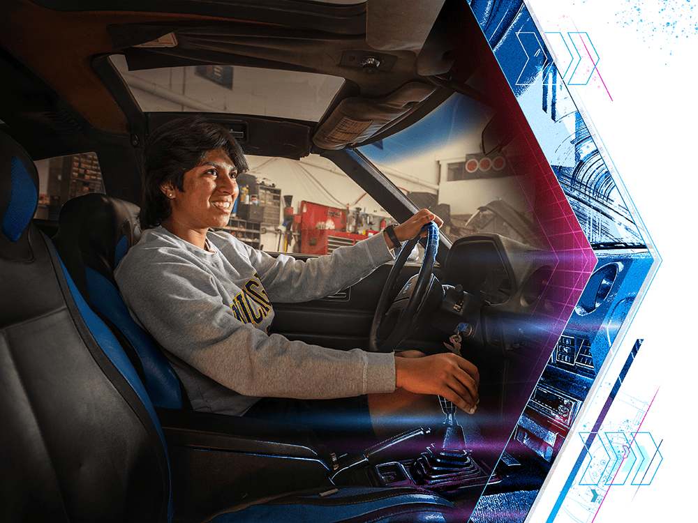 In a photograph, a student wearing a gray University of Michigan crewneck sits in the driver’s seat of a car.