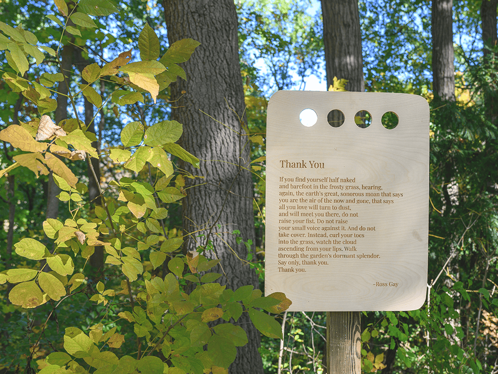 A wooden placard featuring the text of the poem “Thank You,” by Ross Gay, surrounded by trees.