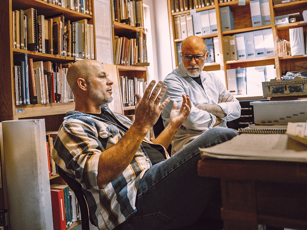 A photograph of two men talking, one in the foreground gesturing and the other with his arms crossed.