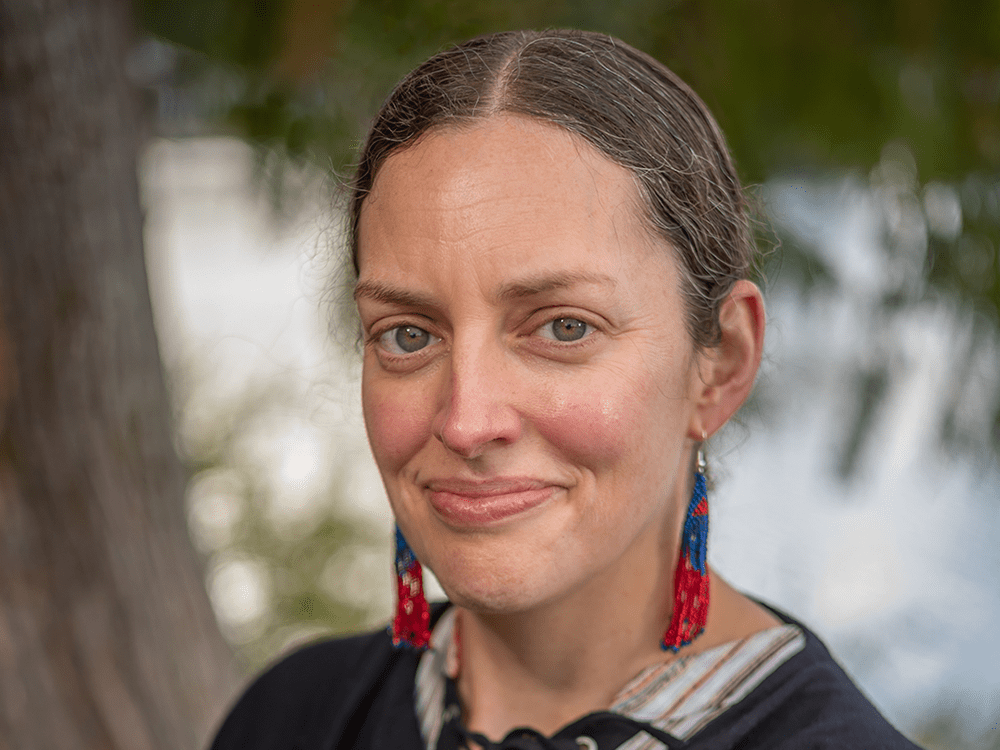 A photograph of a woman looking directly at the camera, wearing beaded earrings and a black blouse.
