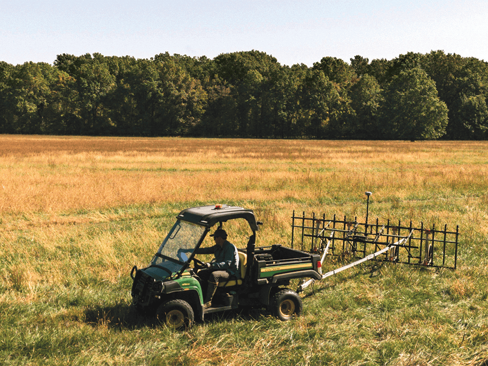 An archaeologist drives a tractor dragging 16 magnetic sensors across a grassy field.