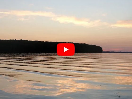 The sun sets off the coast of the calm waters of Douglas Lake as seen from the U-M Biological Station.  A “play” button is visible in the foreground indicating this is a video link.