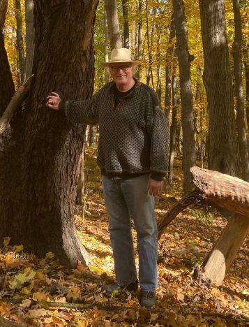 Knute Nadelhoffer stands next to a tree at the forest at the U-M Biological Station. There are fallen yellow leaves on the ground and he's wearing a cowboy-style hat and jeans.