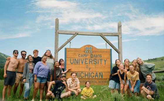 A group of 18 students stand in front of a large wooden sign that displays a maize and blue block M and reads "Camp Davis: University of Michigan."  There are both men and women; they are wearing summer clothes -- shorts, t-shirts, some are shirtless.