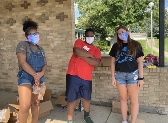Three young people, all wearing protective face masks pose in a group outside a school. The young man in the middle is a middle school camper. The two young women are older, college instructors. All are dressed in shorts for the summer.