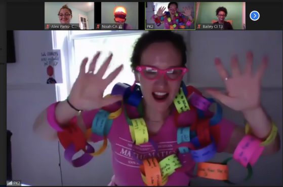 Professor Sarah Koch, wearing pink glasses and t-shirt, is pictured as the featured speaker on a Zoom call with several colleagues. She is wearing a colorful paper chain draped around her shoulders. There are many links to the chain and it is wrapped around her arms and neck several times. You can see that there is writing on each of the links, though you cannot make out the words. She is is doing the "jazz hands" gesture with both hands.