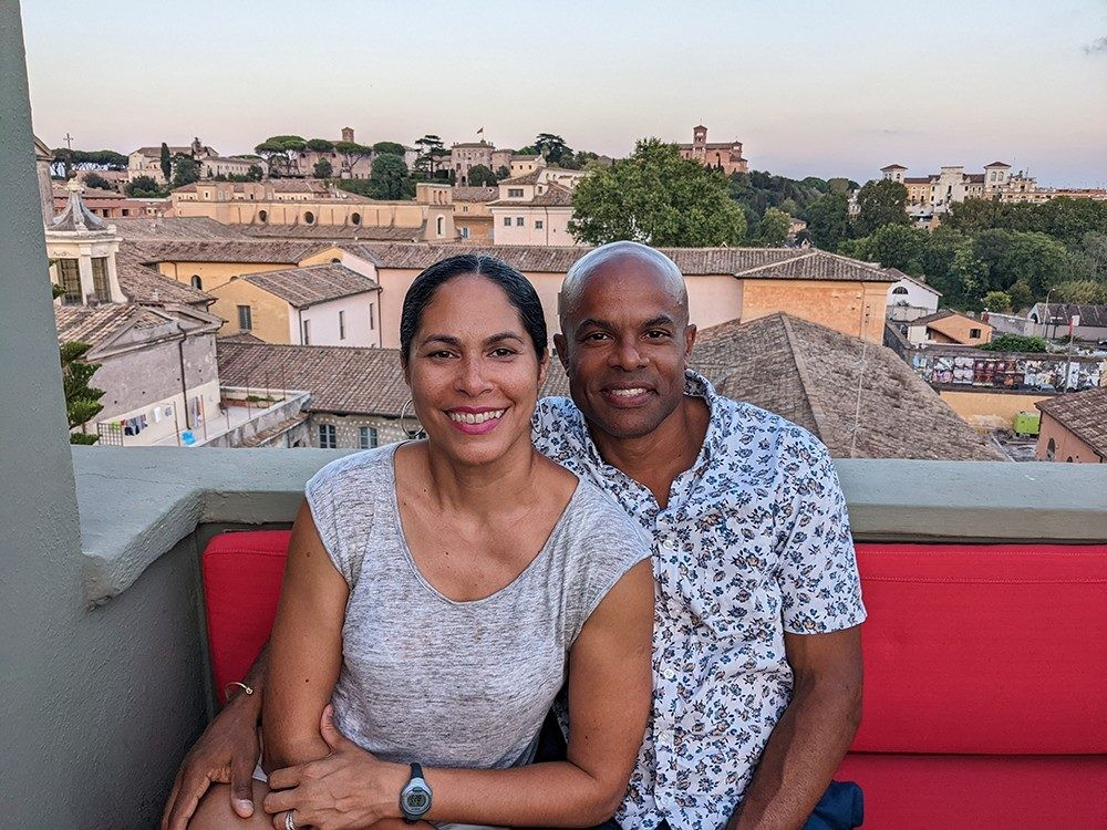 A married couple sits similing on an upholstered red bench on a balcony that overlooks a sunny city.