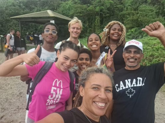 A group selfie of eight people standing on a beach. The are wearing casual clothing, and smiling and waving at the camera.