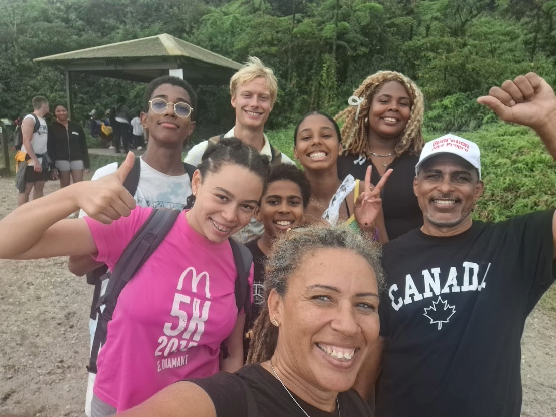 A group selfie of eight people standing on a beach. The are wearing casual clothing, and smiling and waving at the camera.