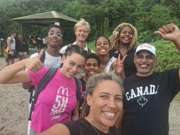 A group selfie of eight people standing on a beach. The are wearing casual clothing, and smiling and waving at the camera.