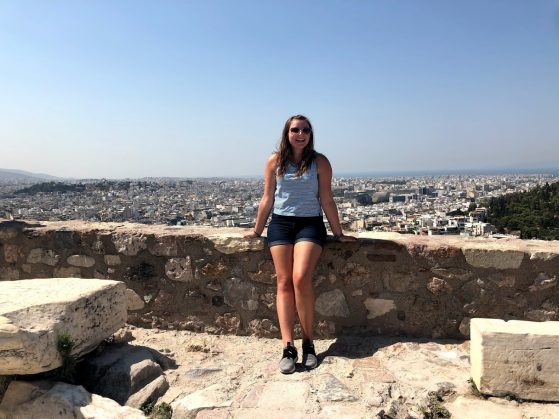 Student in heart-shaped sunglasses, a tank top, and shorts stands atop the Acropolis in Athens, Greece on a beautiful sunny day.
