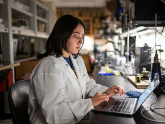 A student clad in a lab coat types on a laptop computer in a university laboratory