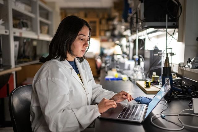 A student clad in a lab coat types on a laptop computer in a university laboratory