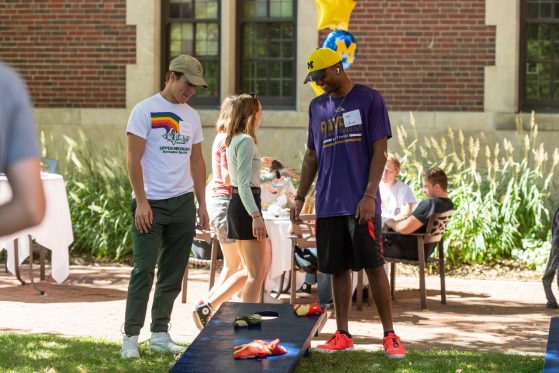 Two male students stand smiling and looking down at a cornhole (bean bag toss) board in the sun-dappled courtyard of the Michigan League. The students are in short sleeves and baseball hats. Other students can be seen in the background, chatting in a group at a round table, and walking in pairs.
