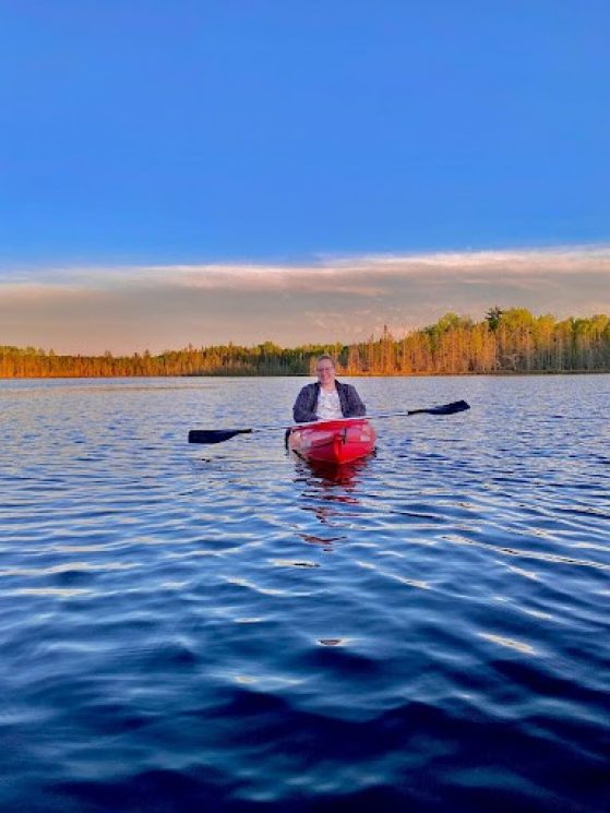 Caitlyn is pictured in a red kayak on a bright blue lake with small ripples. The color of the sky in the background matches the lake. The trees in the background are golden and the clouds are wispy and gauzy.