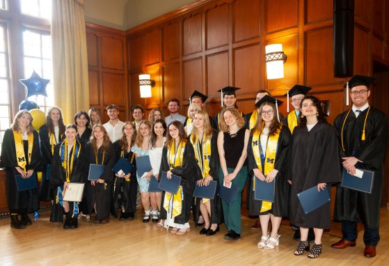 Hapy graduates pose in a group surrounded by the wood floor, wainscotting and a bright window.