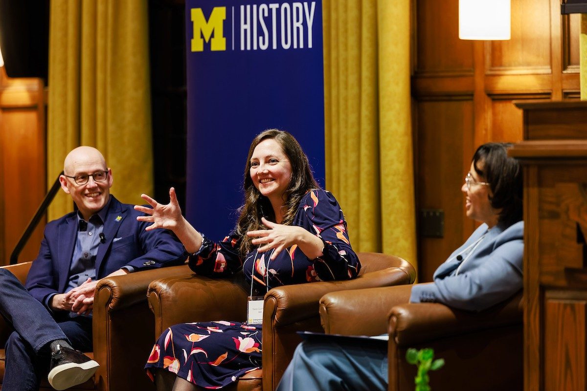 Three people seated on a stage in large brown chairs with a U-M History sign behind them.