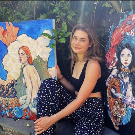 Women with medium-length hair, crop-top and capris, sits between two oils of women painted with bold colors. 