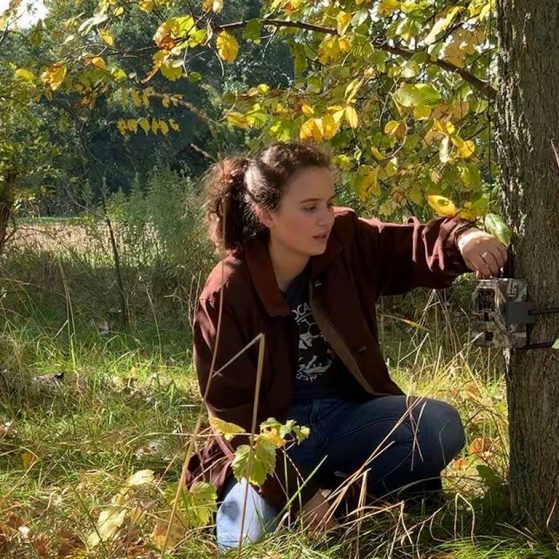 Sam Lima crouches beside lab equipment attached to a tree in an autumn field.