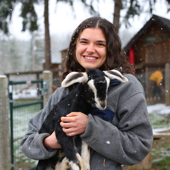 Woman holding small black and white goat. Outdoors, it is snowing. A natural wooden cabin is in the backround.