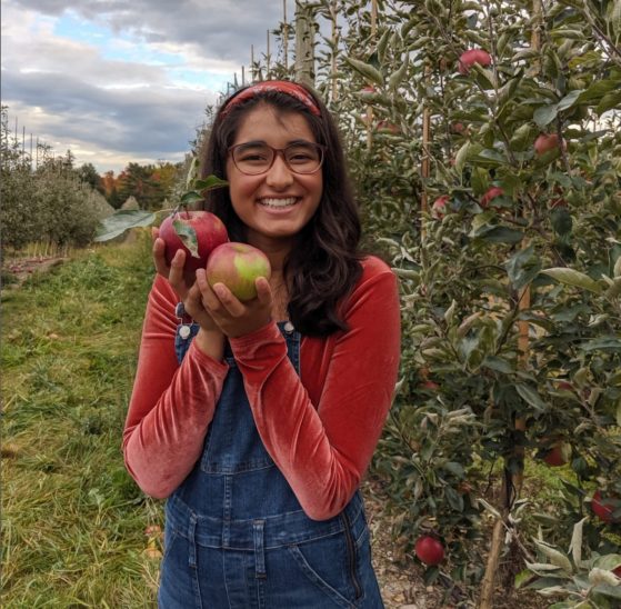 Woman with long hair and a headband stands in an orchard, holding up two apples.