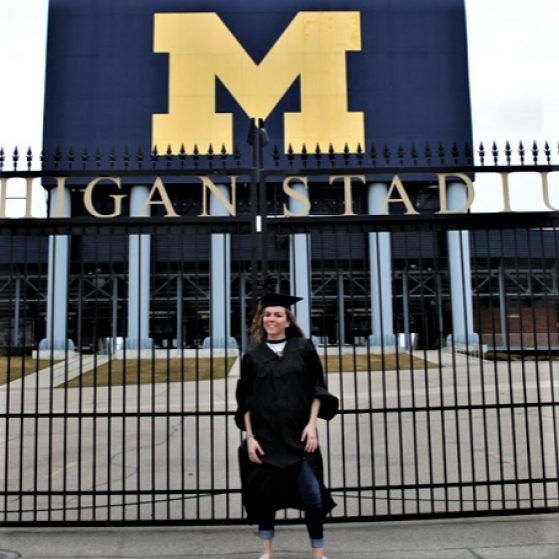 Brooke stands in her cap and gown in front of the Big House block M and stadium gates.