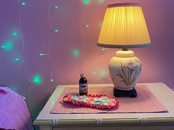 A lamp, eye mask, and small bottle on a bedside table with turquoise string lights hung on the wall.