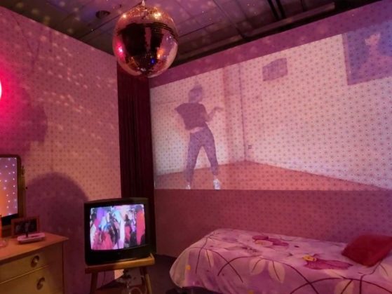 A box tv, bed, disco ball, and video projection of a person dancing fill a pink lit gallery space.