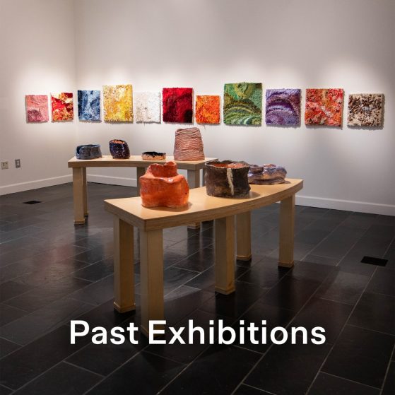 A series of ceramic vessels rest atop two curved wooden tables, with colorful square textile wall works in the background.