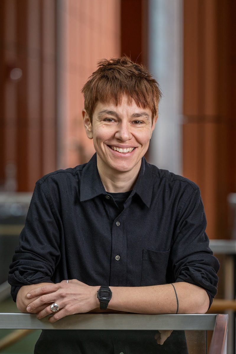 Woman with short red hair wearing a black button-up shirt leans on a railing with her arms crossed smiling for a professional headshot
