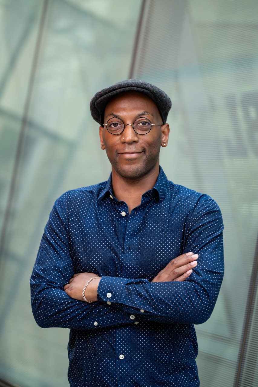 Man wearing glasses, gray cabby hat, and blue button up shirt poses smiling with arms crossed for a professional headshot