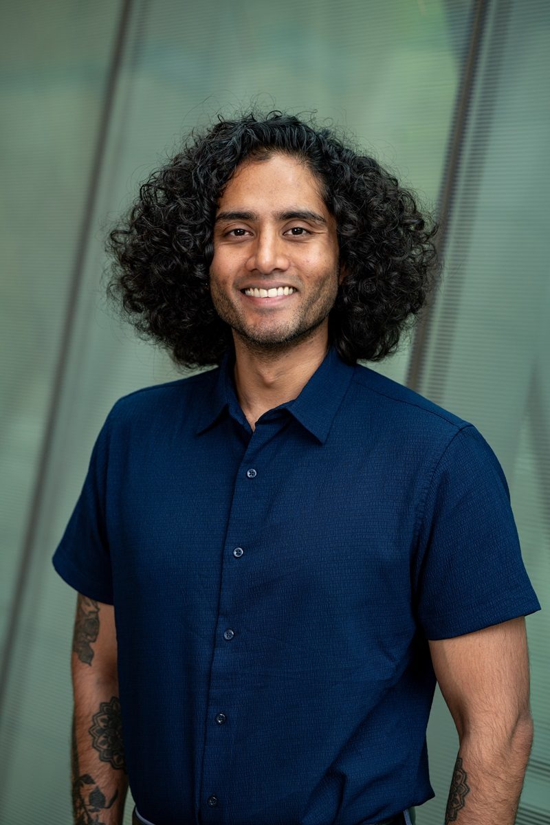 Man with long black curly hair and a shortsleeve blue button up smiles widely at the camera for a professional headshot