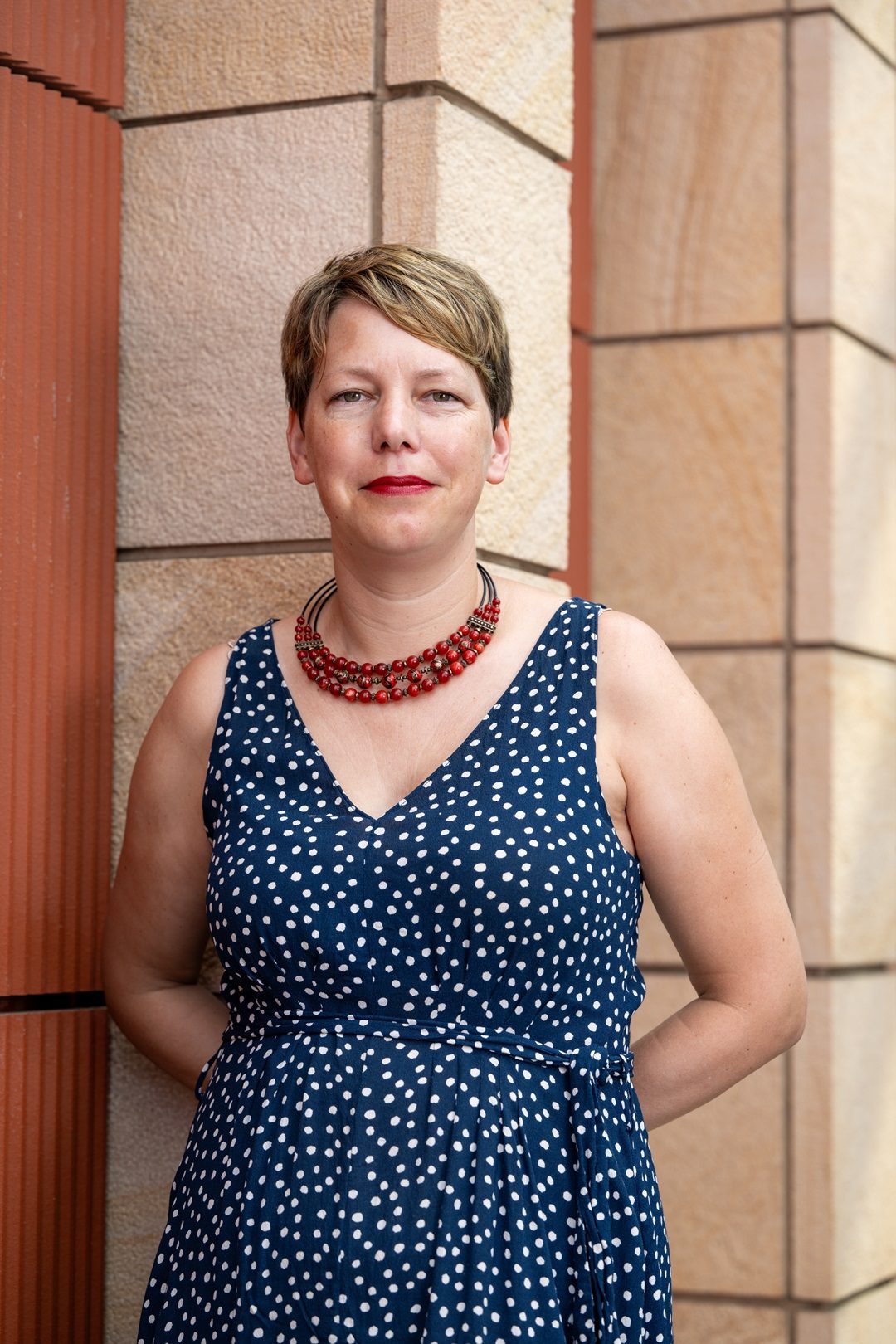 A woman with short blond hair in a blue dress poses with her hands behind her back for a professional headshot