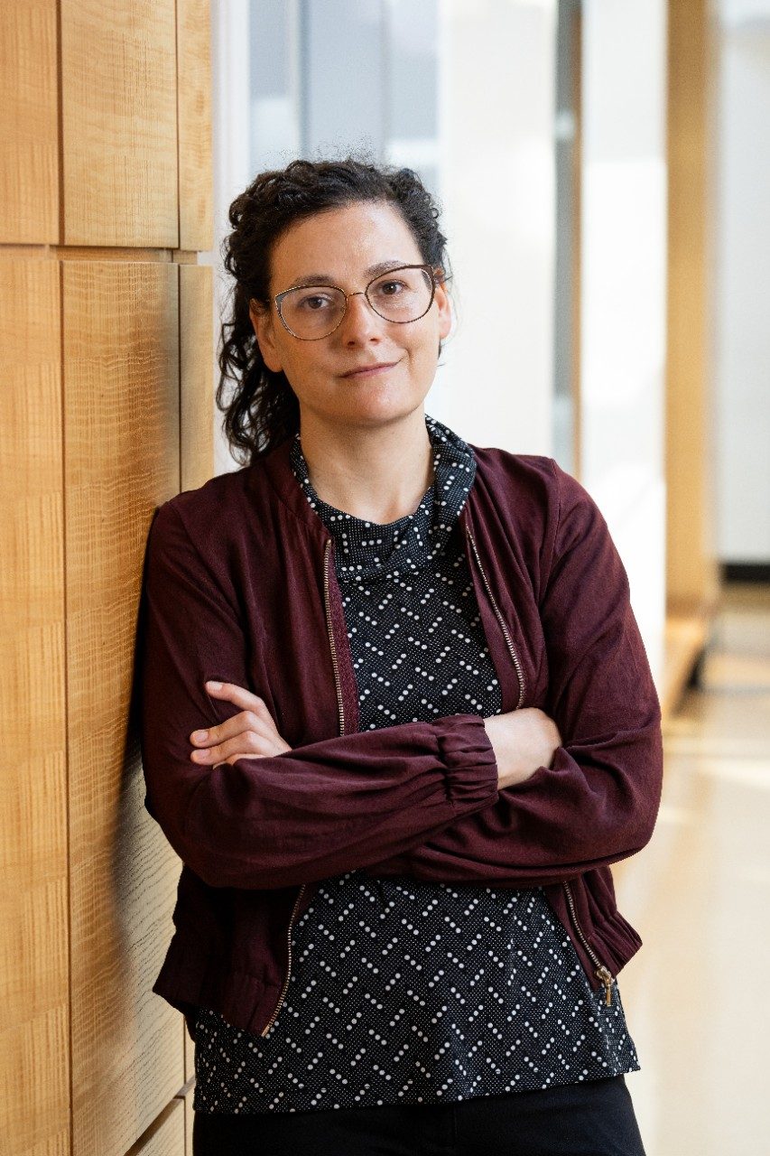 A woman with curly brown hair wearing a black blouse and red cardigan poses smiling with her arms crossed for a professional headshot