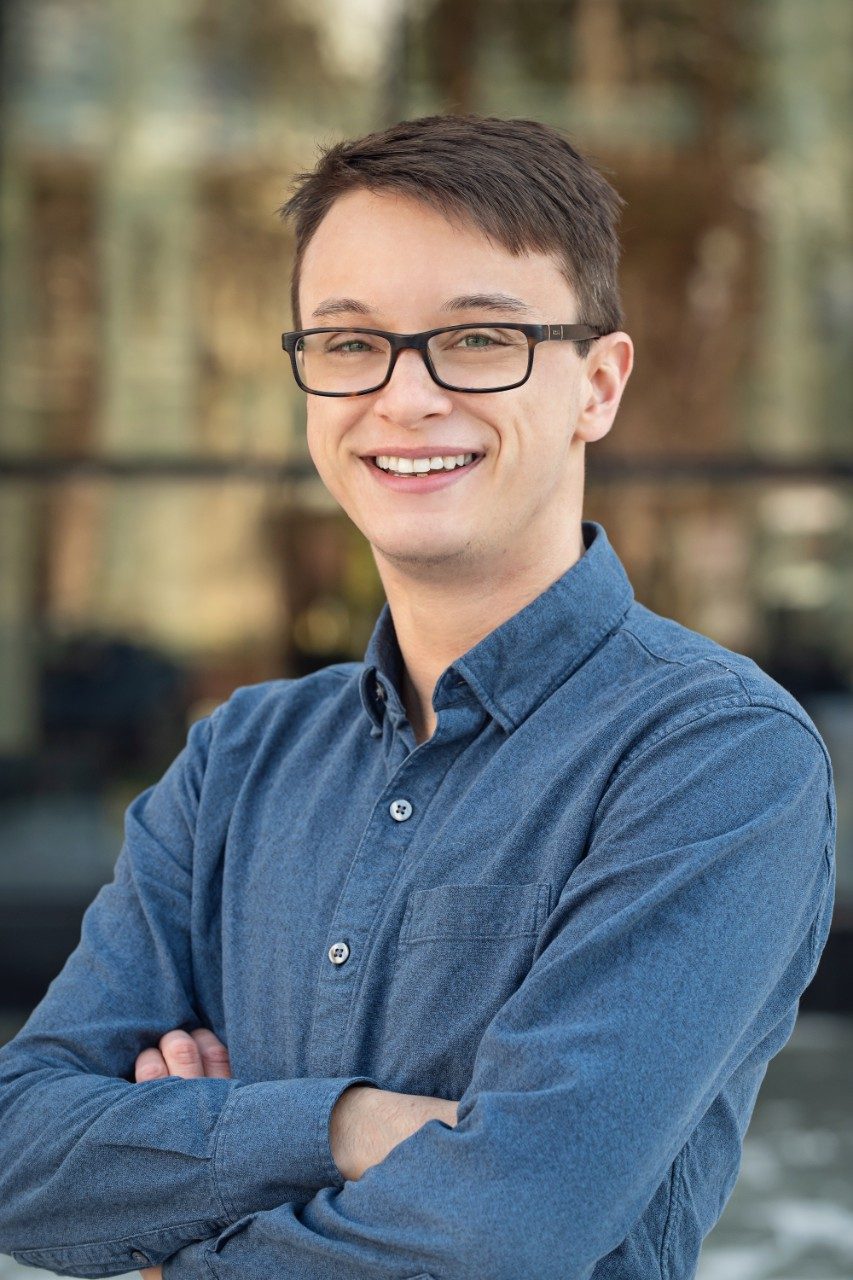 a man with short brown hair and glasses poses smiling with his arms crossed for a professional headshot