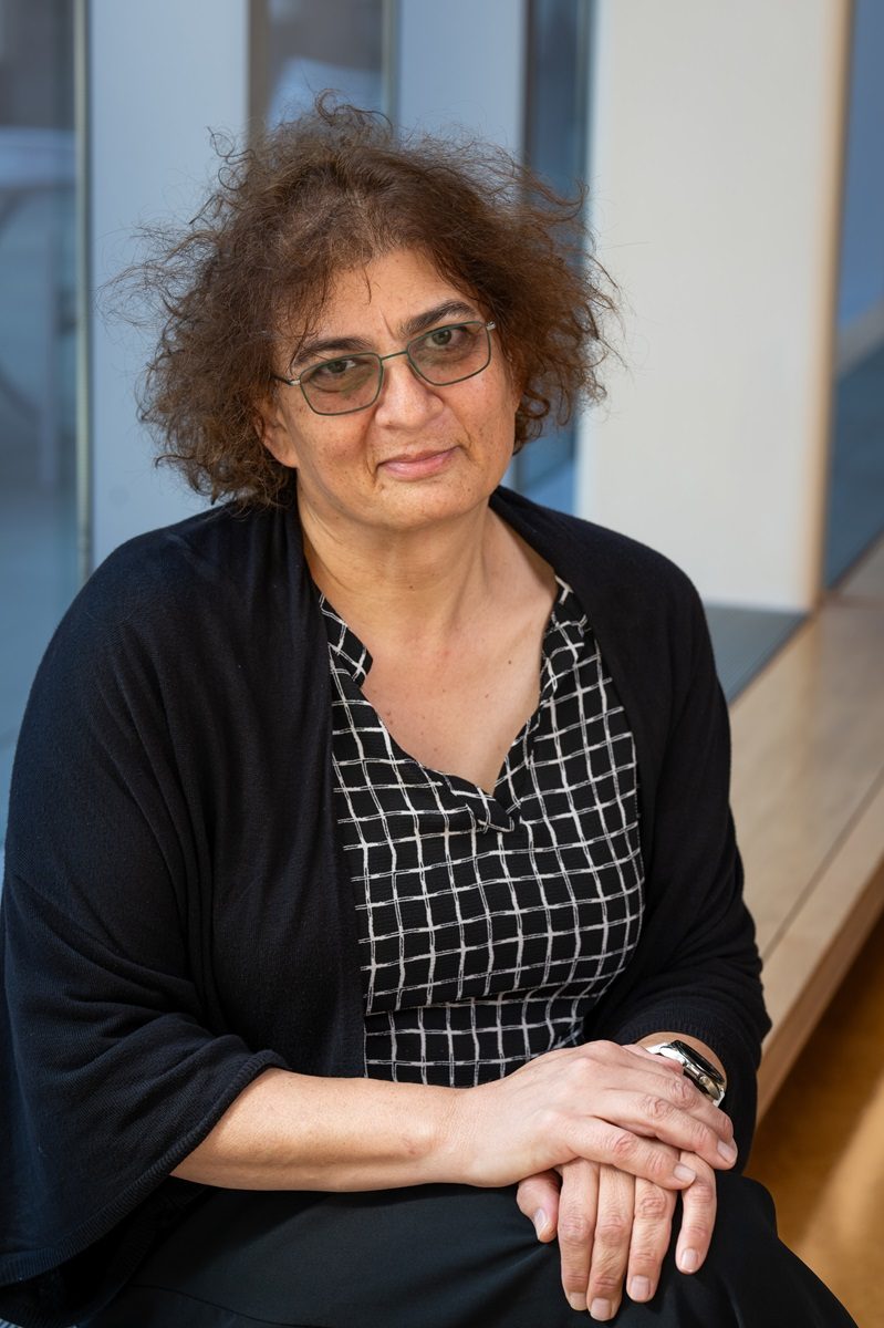 Woman with curly hair and glasses wearing a black and white patterned blouse under a black cardigan smiles at the camera for a professional headshot