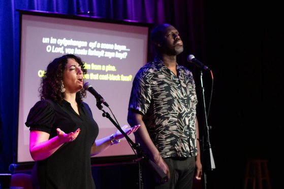 short white lady with curly brown hair and tall bald black man sing on stage side by side