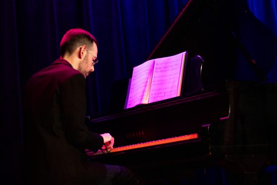 Skinny white man with short hair playing piano under purple lights