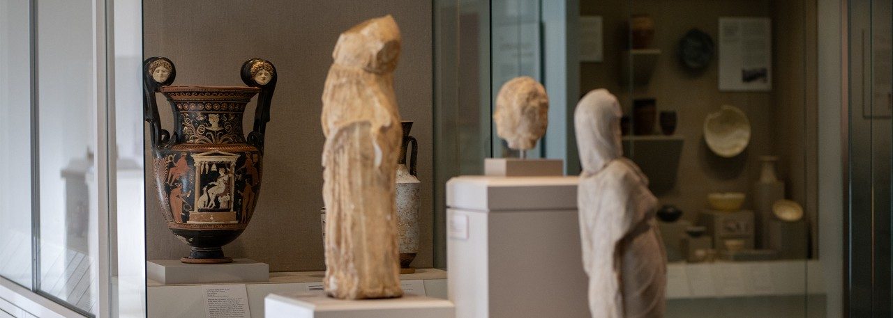 View of the Kelsey Museum’s Greek galleries, showing three white marble statues displayed on podiums and a large two-handled amphora in a case behind them.