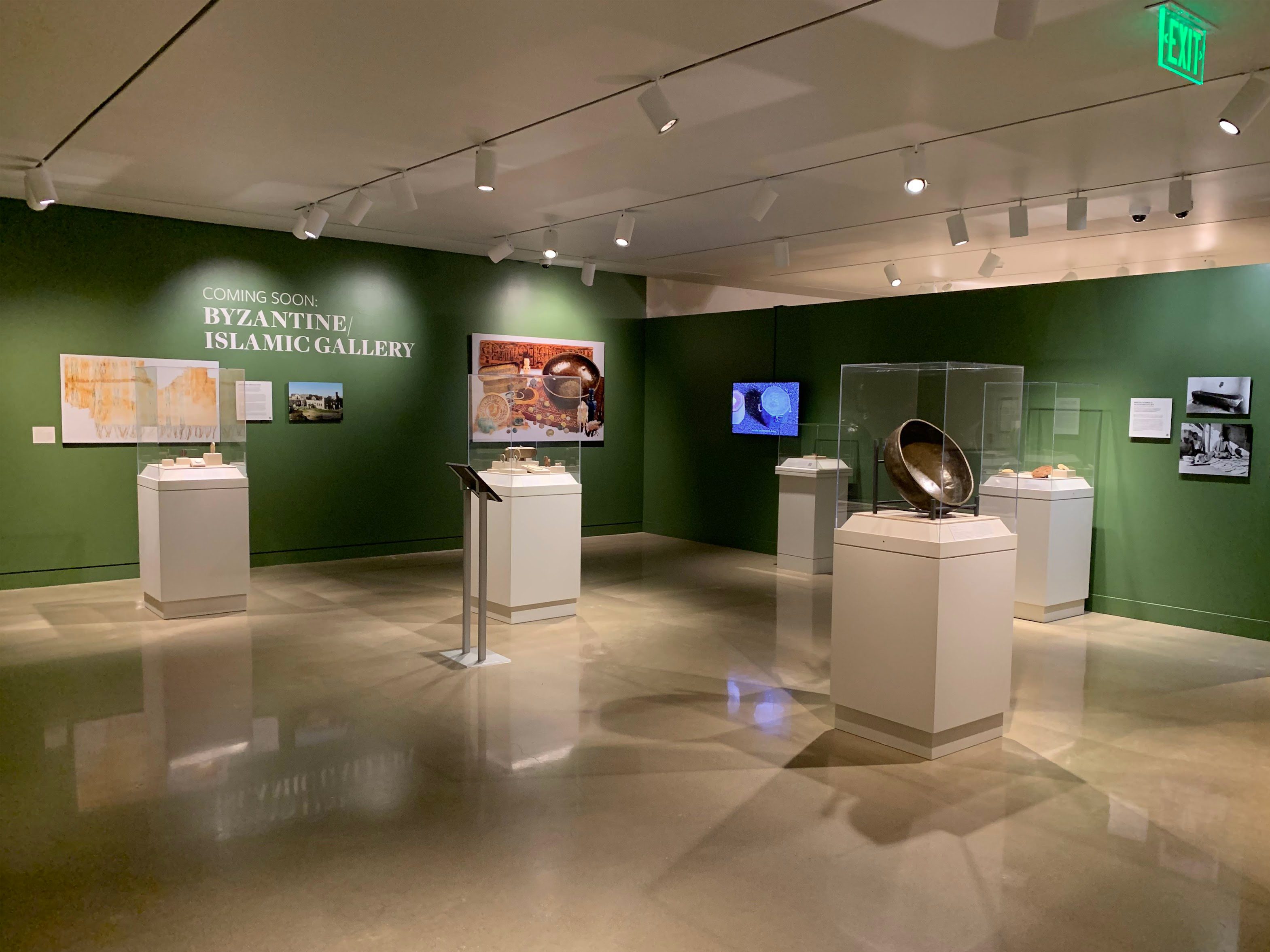 Museum gallery space with five vitrines containing artifacts. Hanging on the walls are photos, including that of a textile and an assemblage of artifacts, as well as a TV showing a video. Text on the wall reads, “Coming Soon: Byzantine/Islamic Gallery.”