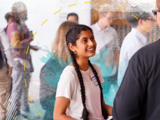 Female student with long black hair in two braids smiles while speaking to an employer at a career fair event in the LSA building.