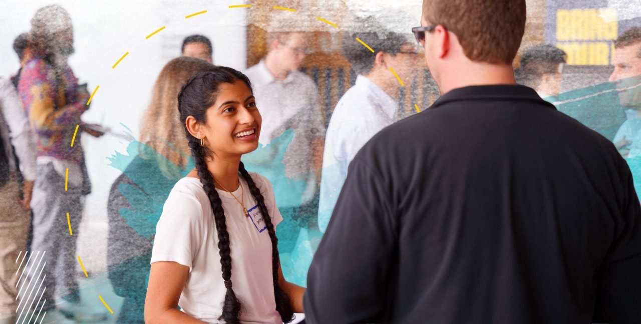 Female student with long black hair in two braids smiles while speaking to an employer at a career fair event in the LSA building.