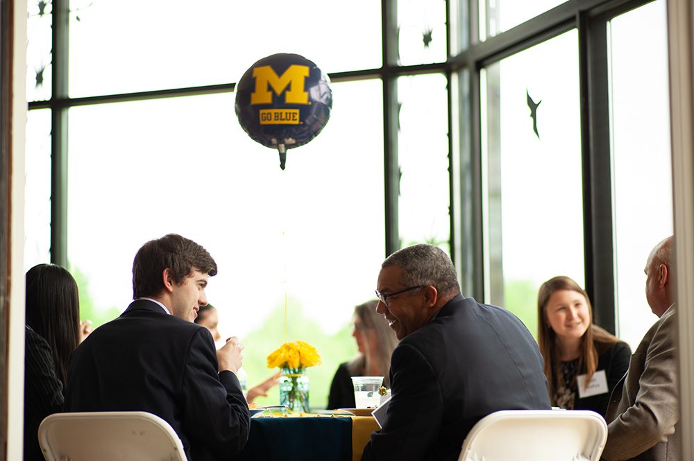 An alum and student seated at a table, with their backs to the camera; they are talking over lunch and smiling. A blue U-M balloon floats above them.
