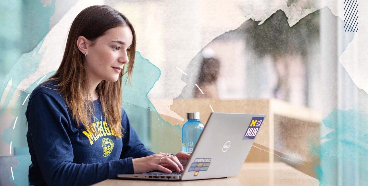 A person wearing a Michigan sweatshirt works on a laptop with LSA Hub stickers, seated at a table with a blue water bottle.