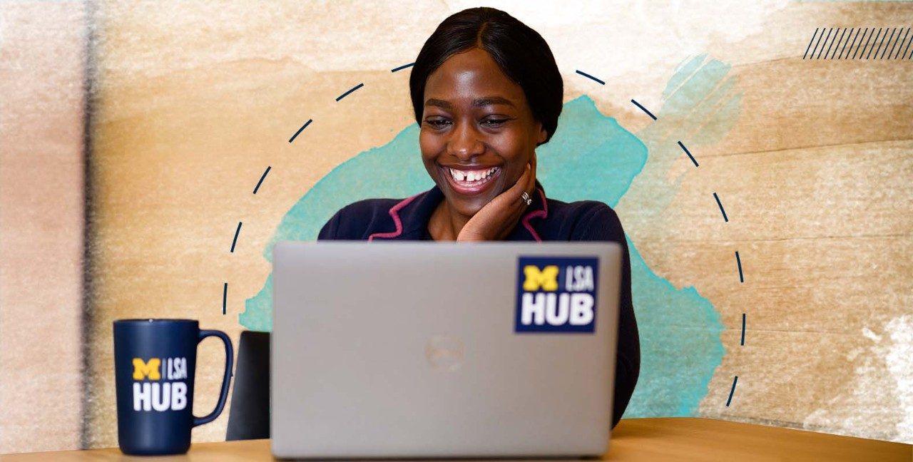 Student smiling while looking at their laptop. They're resting their head on their hand and has a LSA Hub mug off to one side on the table they're working on.