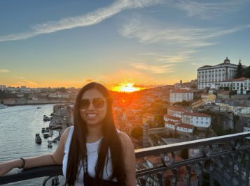 Aashna stands outdoors against a railing overlooking a canal and coastal city at sunset
