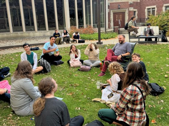 group of students sitting on the grass while their male professor holds class from a lawn chair